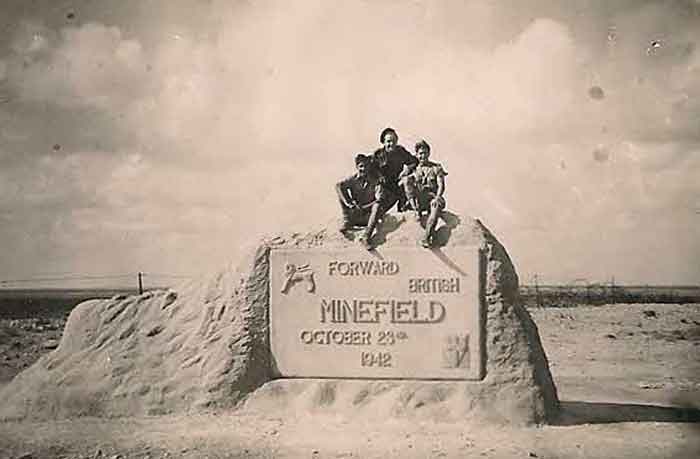 large stone monument with 'forward british minefield, october 23rd, 1942' engraved on it. three men have climbed on top.